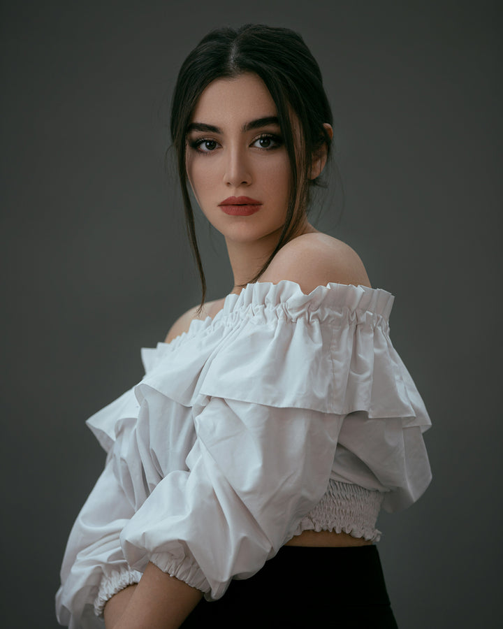 Woman with dark hair looking toward the camera in a white off-the-shoulder blouse against a dark background.