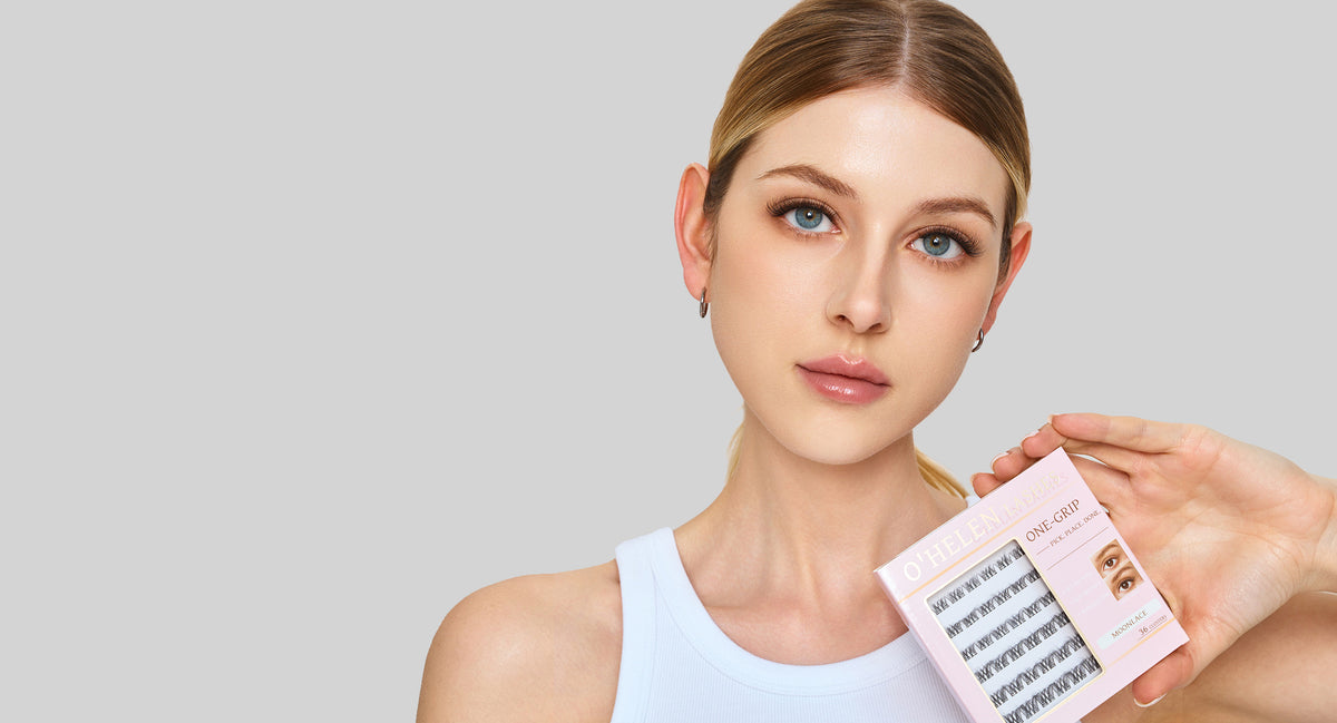 Wide banner showing a woman with blue eyes in a white top holding a pink O’HELEN One-Grip lash pack against a gray background.
