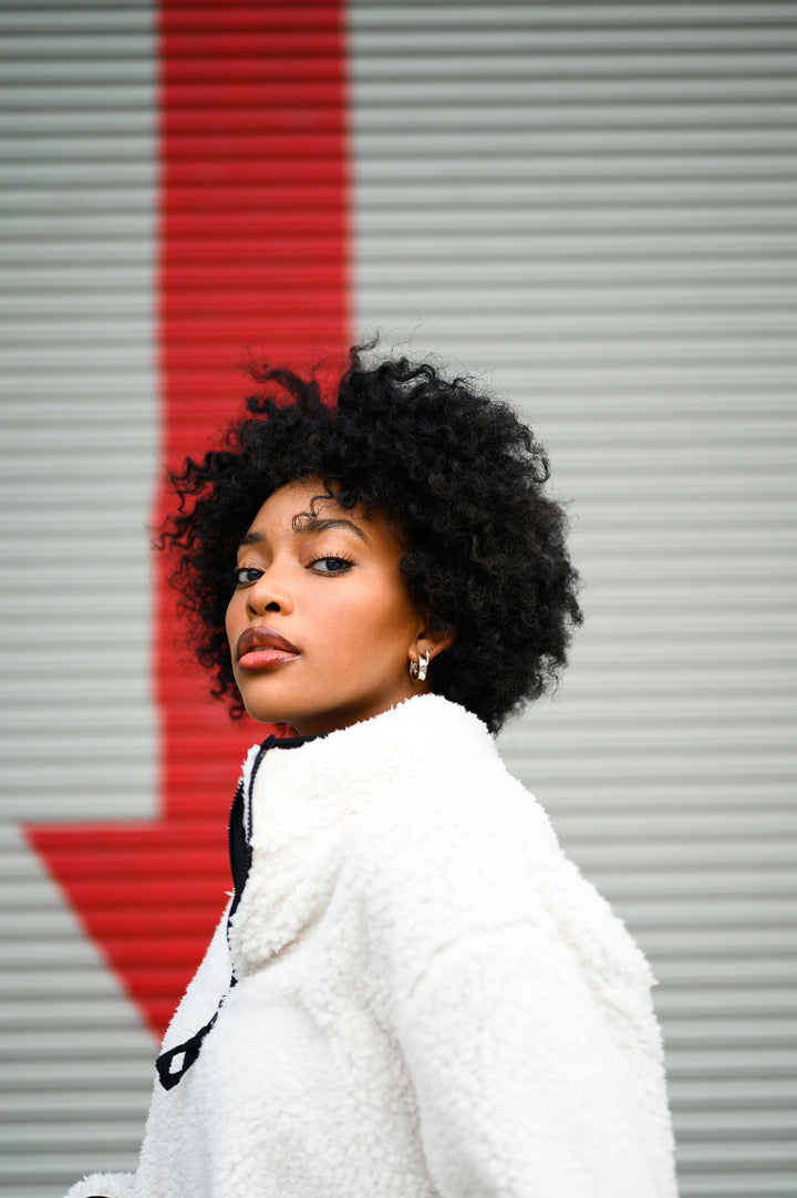 Woman with short curly hair in a white jacket looking back toward the camera in front of a gray shutter with a red stripe.