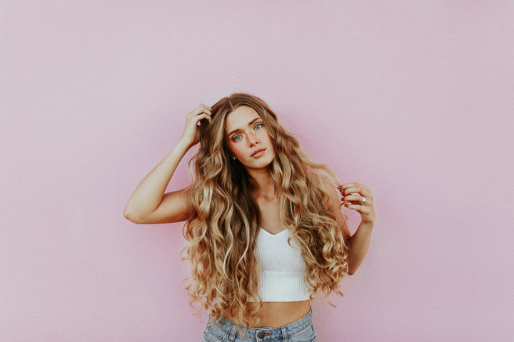 Woman with long wavy hair wearing a white top and holding her hair against a pink background.